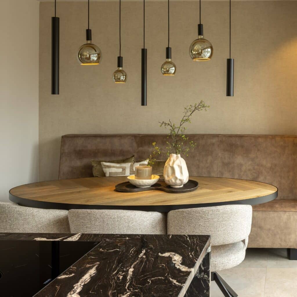 Moderne leefruimte, voorzien van zithoek met ovale houten tafel, bank en drie stoelen. We kijken ernaar vanuit een moderne keuken met zwart marmerlook kookeiland.